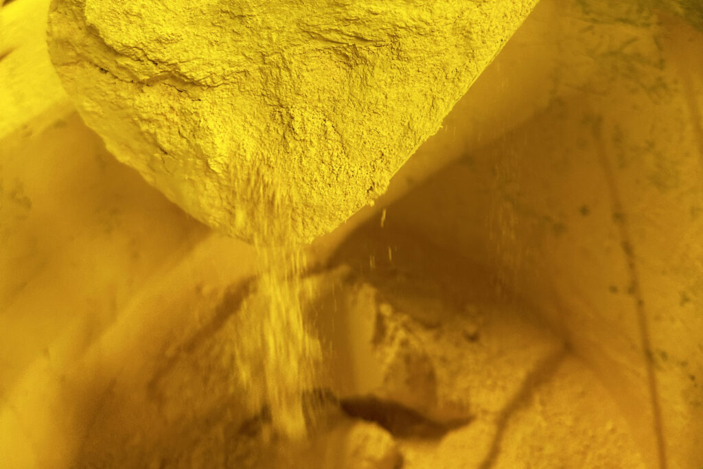 Ein gelbes Pigment rieselt von einer Schaufel voll davon zurück in die große Tonne.