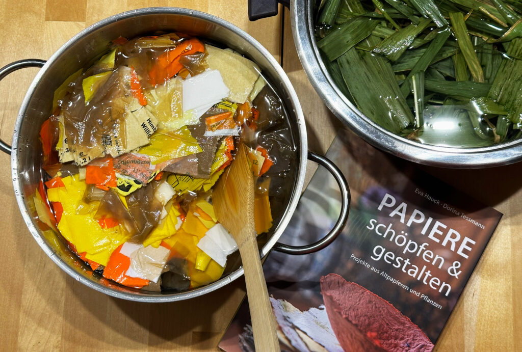 Zwei Töpfe mit eingeweichten bunten Papierschnipseln und Maisblättern sollen zu Pulpe werden, wie in dem Buch "PApiere schöpfen und gestalten", das daneben liegt, erklärt wird.