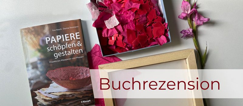 Das Buch "Papiere schöpfen und gestalten" liegt auf dem Tisch neben pinken Papierschnipseln, dem Schöpfrahmen und einer violetten Blüte.