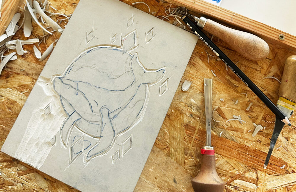 Linolplatte mit aufgebrachter Skizze und Schnitzwerkzeug auf einem Holzbrett.