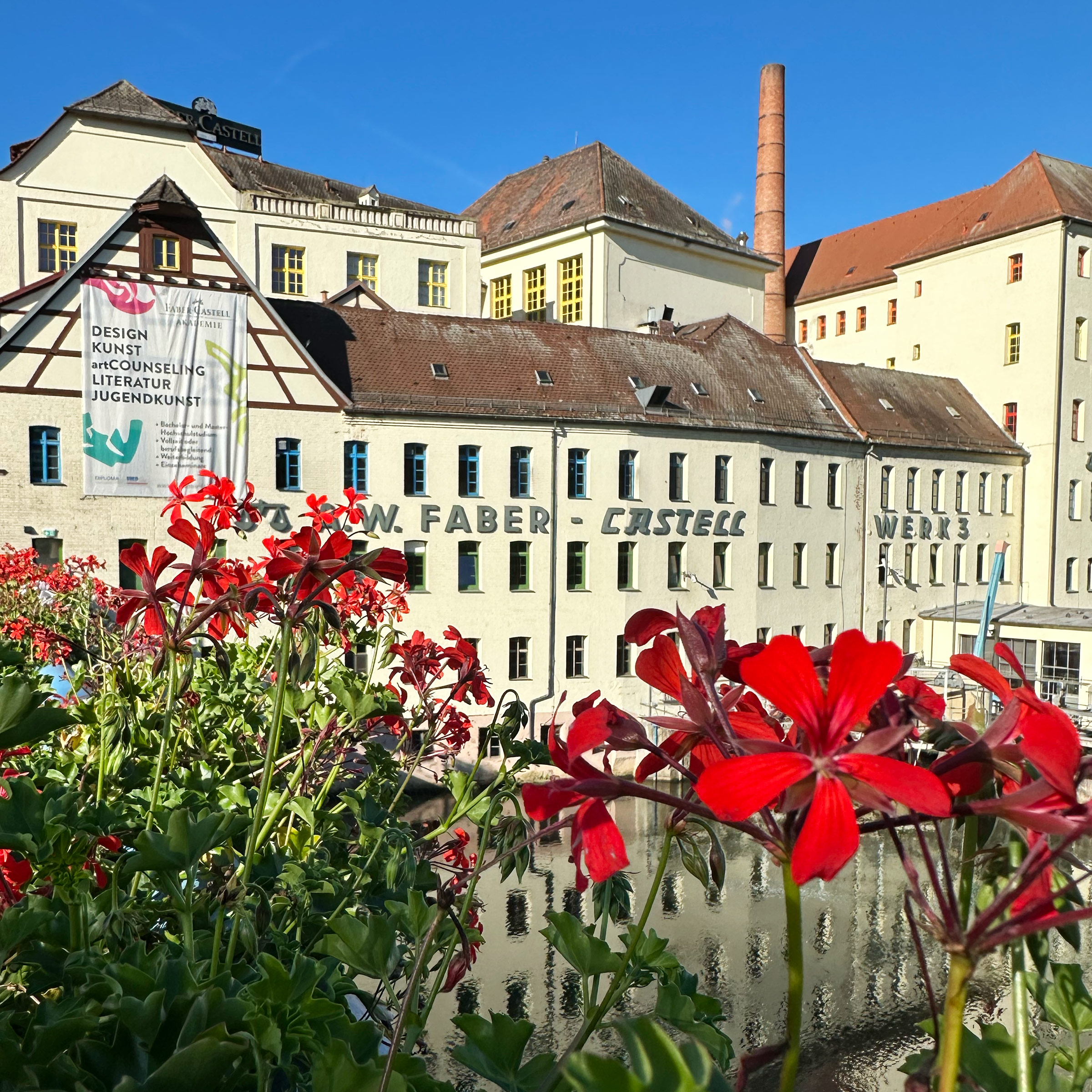 Das Werk 3 beheimtat die Akademie Faber-Castell, direkt an der Rednitz gelegen, mit den typischen Farben der Fensterrahmen je nach Stockwerk. Im Vordergrund sind rote Geranien am Brückengeländer zu sehen.