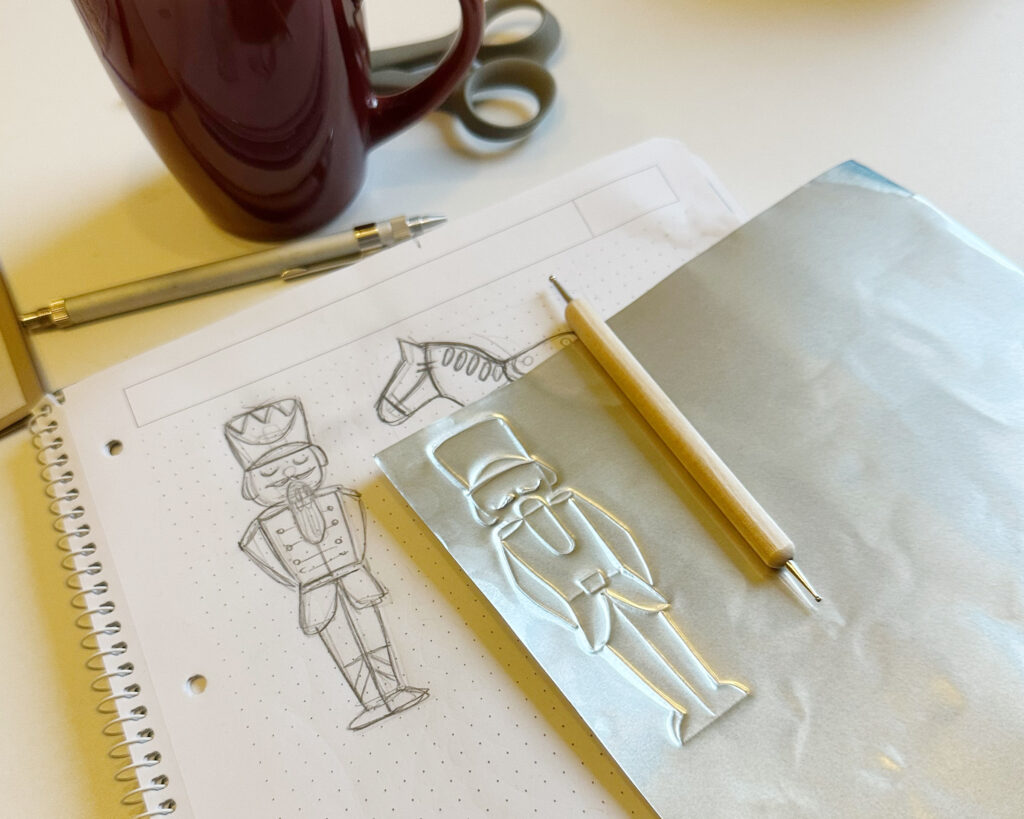 Die Skizze auf dem College-Block, der gleichzeitig auch als weiche Unterlage dient, neben der silbernen Prägefolie mit den ersten nachgefahrenen Linien des Nussknackers und dem Prägestift. Im Hintergrund eine Schere und eine Teetasse.